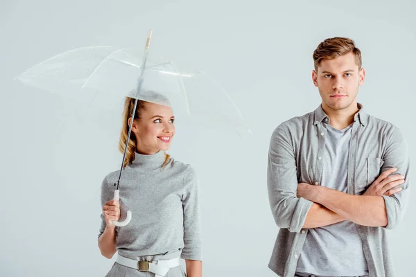 Donna sorridente con ombrello trasparente e in piedi vicino all'uomo insoddisfatto con le braccia incrociate isolate sul grigio — Foto stock