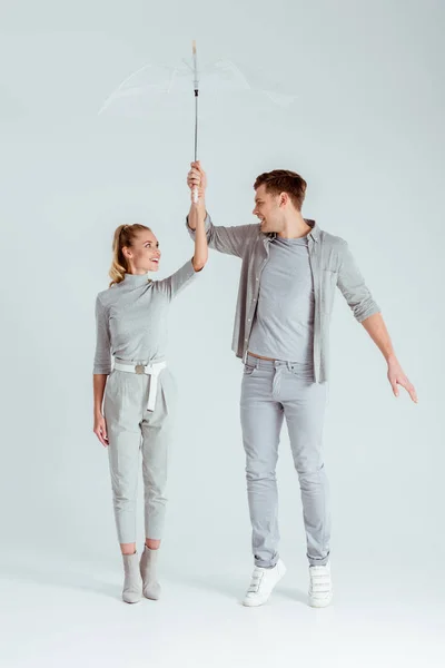 Happy couple looking at each other, standing on tiptoe and posing with transparent umbrella on grey background — Stock Photo