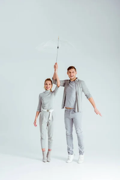 Glückliches Paar blickt in die Kamera, steht auf Zehenspitzen und posiert mit transparentem Regenschirm auf grauem Hintergrund — Stockfoto
