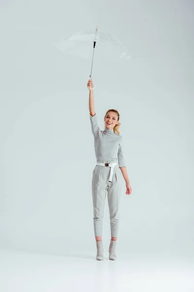 Schöne Frau blickt in die Kamera, steht auf Zehenspitzen und posiert mit transparentem Regenschirm auf grauem Hintergrund — Stockfoto