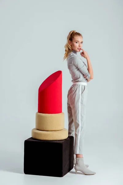 Hermosa mujer mordiendo dedo y posando cerca de gran modelo de lápiz labial rojo sobre fondo gris - foto de stock