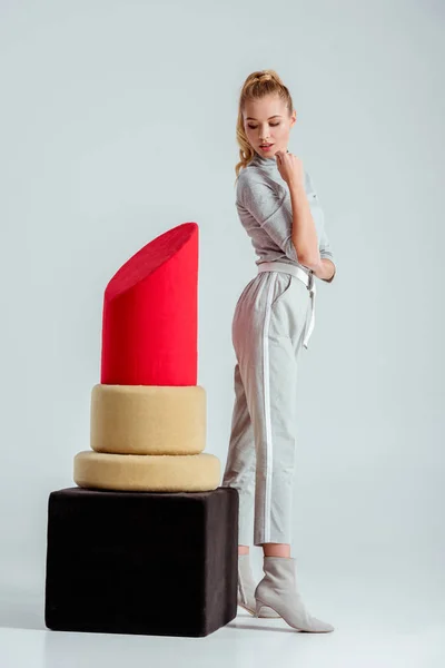 Beautiful woman looking down and posing near big red lipstick model on grey background — Stock Photo