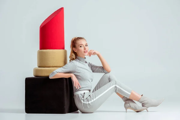 Beautiful woman touching chin and posing near big red lipstick model on grey background — Stock Photo