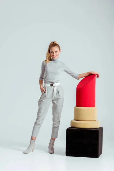 Mujer sonriente mirando a la cámara y posando cerca de un gran modelo de lápiz labial rojo sobre fondo gris - foto de stock