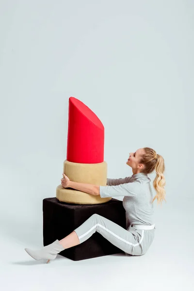 Femme assise et tenant un grand modèle de rouge à lèvres rouge sur fond gris — Photo de stock