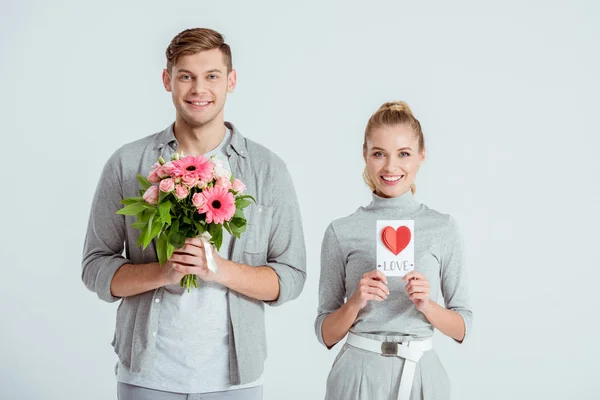 Happy couple looking at camera and holding Valentines card with flower bouquet isolated on grey — Stock Photo