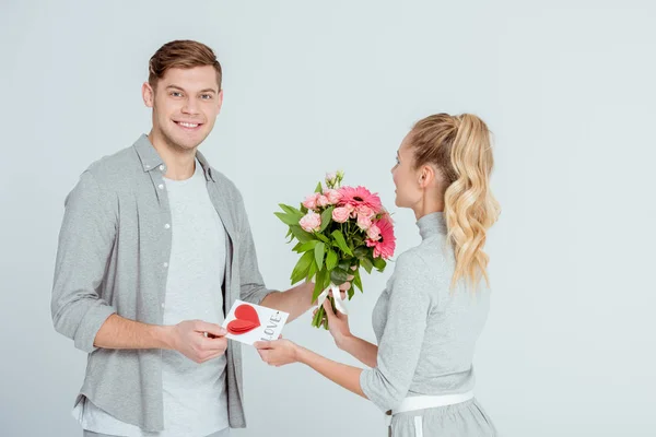 Homem sorridente saudação mulher com cartão de Valentim e buquê de flores isolado em cinza — Fotografia de Stock