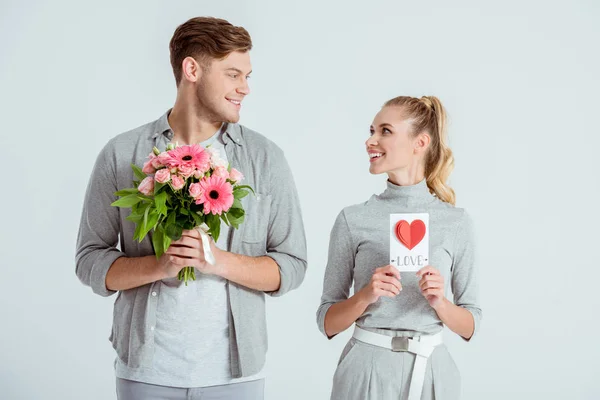 Happy couple looking at each other and holding Valentines card and flower bouquet isolated on grey — Stock Photo