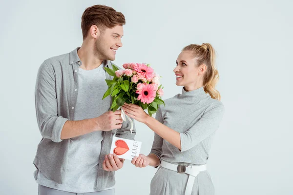 Красивий чоловік вітає жінку з листівкою Валентина і квітковим букетом ізольовано на сірому — стокове фото