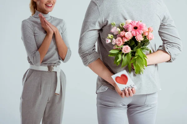 Cropped view of woman with folded hands looking at man hiding flower bouquet and Valentines greeting card behind back isolated on grey — Stock Photo