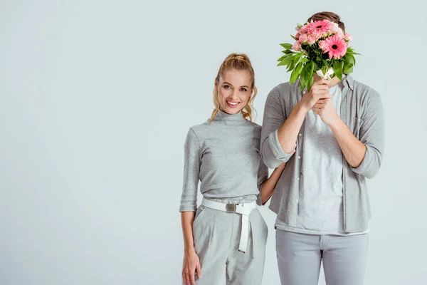Woman looking at camera and standing near man hiding behind flower bouquet isolated on grey — Stock Photo