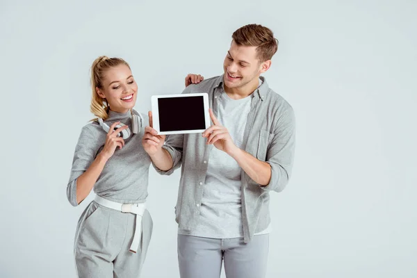 Улыбающаяся женщина в наушниках, стоящая рядом с мужчиной, держащая цифровой планшет с пустым экраном, изолированным на сером — стоковое фото
