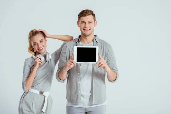 Schöne Frau mit Kopfhörern, die neben einem Mann steht und ein digitales Tablet mit leerem Bildschirm präsentiert, isoliert auf grau — Stockfoto