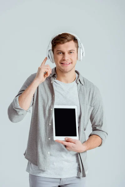 Hombre guapo apuntando con el dedo a los auriculares mientras sostiene la tableta digital con pantalla en blanco aislado en gris — Stock Photo