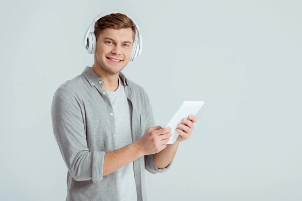 Mann mit Kopfhörer hört Musik, blickt auf Kamera und benutzt digitales Tablet isoliert auf grau — Stockfoto