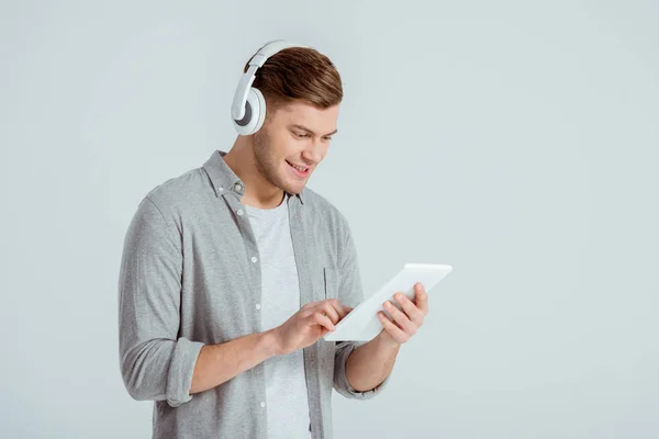 Homem bonito em fones de ouvido ouvindo música e usando tablet digital isolado em cinza — Fotografia de Stock