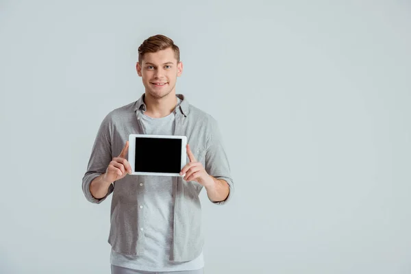 Hombre guapo en ropa gris mirando a la cámara y presentando tableta digital con pantalla en blanco aislado en gris - foto de stock