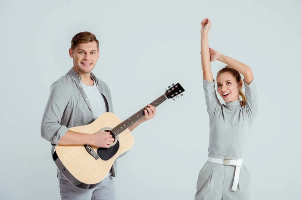 Schöner Mann spielt Akustikgitarre, während schöne Frau isoliert auf grau tanzt — Stockfoto