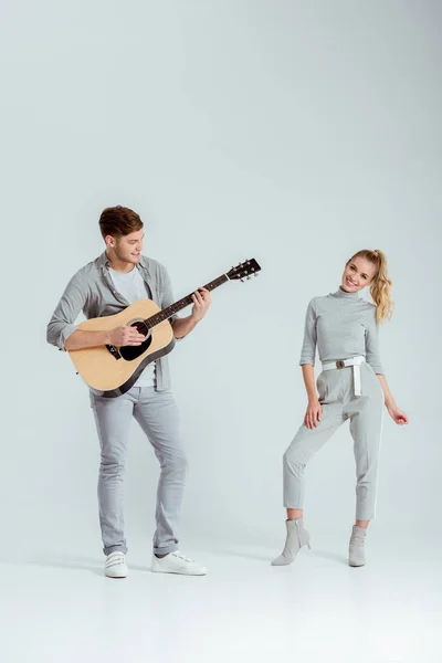 Мужчина играет на акустической гитаре, а улыбающаяся женщина танцует на сером фоне — стоковое фото
