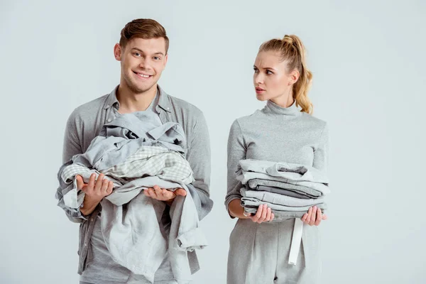 Dissatisfied woman looking at smiling man holding grey clothes isolated on grey — Stock Photo