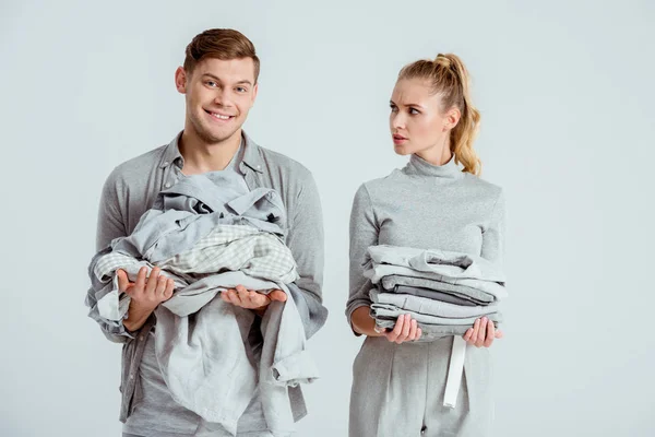 Donna insoddisfatta guardando l'uomo sorridente che tiene vestiti grigi isolati su grigio — Foto stock