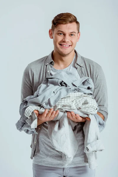 Hombre guapo sonriente mirando a la cámara y sosteniendo una pila de ropa gris aislada en gris - foto de stock