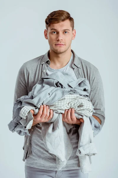 Hombre guapo mirando a la cámara y sosteniendo una pila de ropa gris aislada en gris - foto de stock