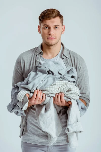 Bello uomo guardando la macchina fotografica e tenendo mucchio di vestiti grigi isolati su grigio — Foto stock