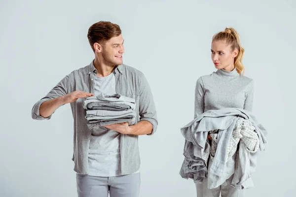 Donna insoddisfatta guardando l'uomo sorridente che tiene vestiti grigi isolati su grigio — Foto stock