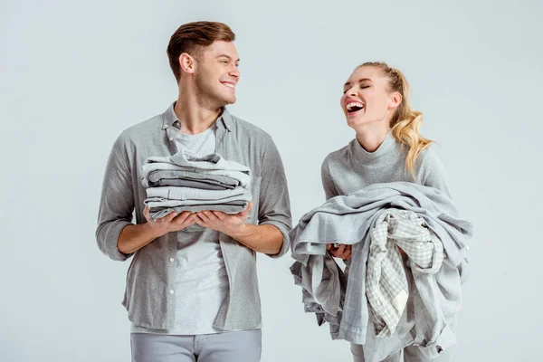 Belo casal segurando pilhas de roupas cinza e rindo isolado em cinza — Fotografia de Stock