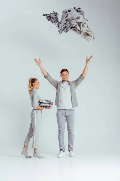 Woman in grey outfit standing near man throwing pile of clothes in air on grey background — Stock Photo