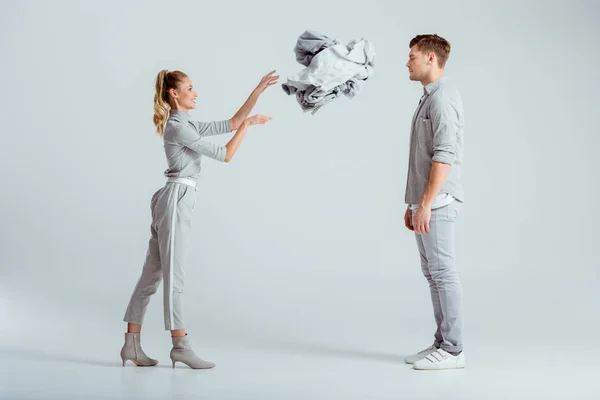 Smiling woman throwing pile of clothes at man on grey background — Stock Photo