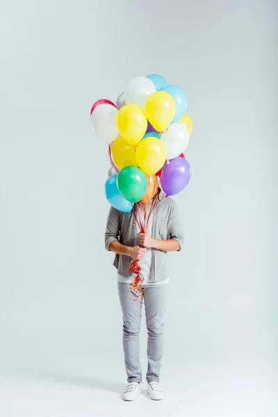 Man in grey clothes hiding behind bundle of colorful balloons on grey background — Stock Photo