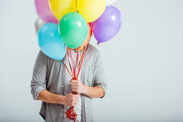 Hombre en ropa gris escondido detrás de manojo de globos de colores con espacio de copia sobre fondo gris - foto de stock