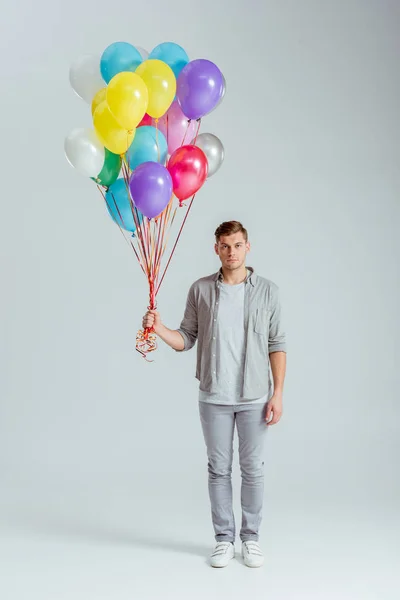 Man in grey clothing holding bundle of colorful balloons on grey background — Stock Photo