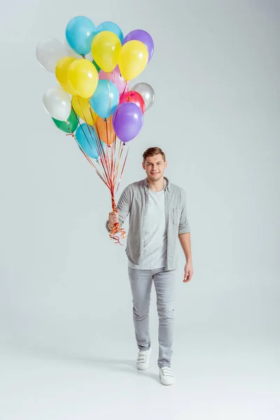 Handsome man holding bundle of colorful balloons and looking at camera on grey background — Stock Photo