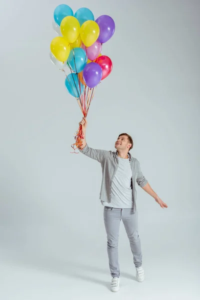 Happy man in grey clothing holding bundle of colorful balloons on grey background — Stock Photo