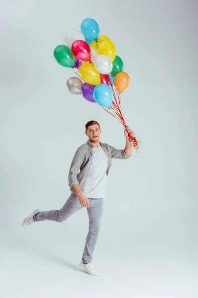 Mann springt mit Bündel bunter Luftballons und blickt in Kamera auf grauem Hintergrund — Stockfoto