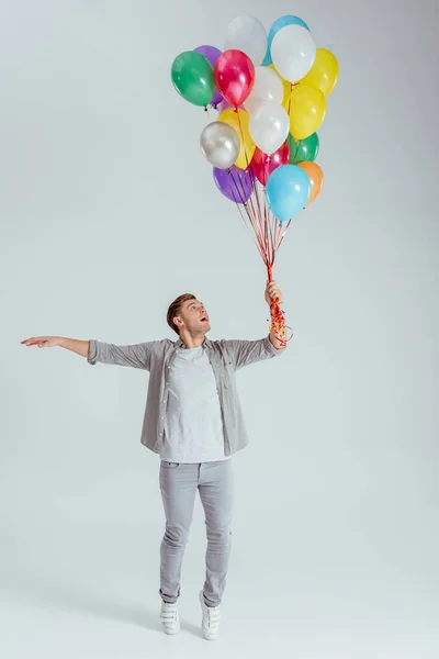 Man in grey clothing standing on tiptoe and holding bundle of colorful balloons on grey background — Stock Photo