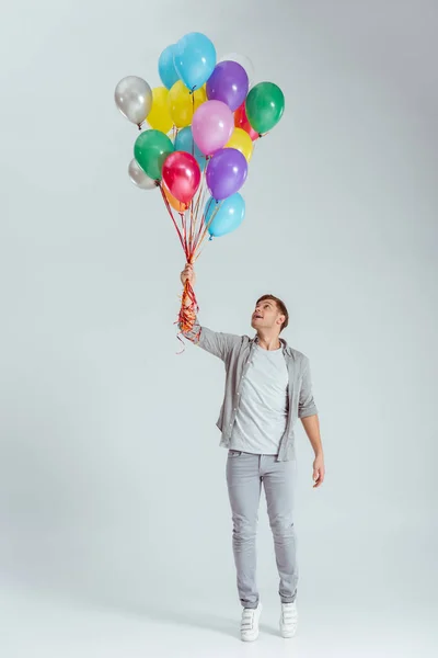 Handsome man standing on tiptoe and holding bundle of colorful balloons on grey background — Stock Photo