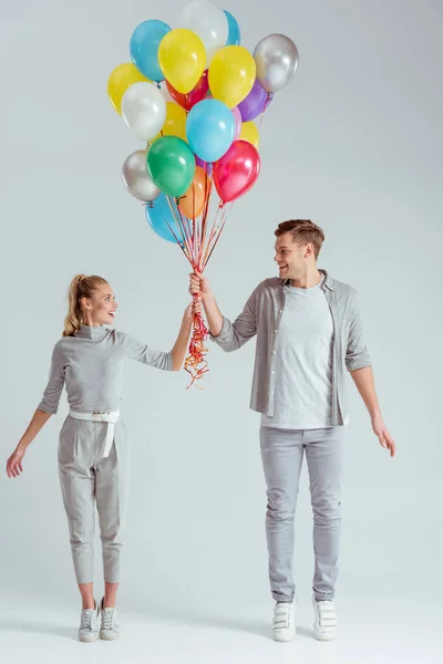 Happy couple standing on tiptoe and holding bundle of colorful balloons on grey background — Stock Photo