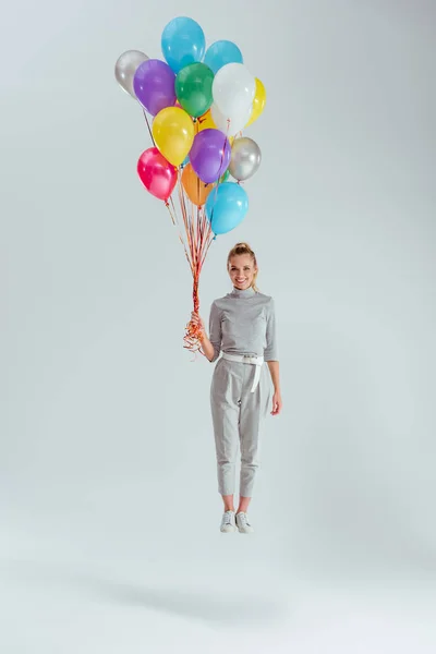 Femme souriante regardant la caméra et sautant dans l'air avec un paquet de ballons colorés sur fond gris — Photo de stock