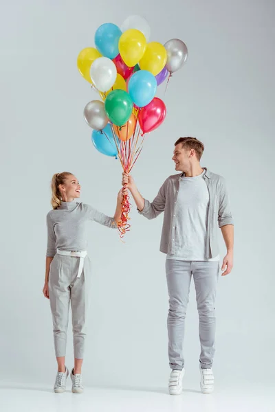 Pareja de pie de puntillas, mirándose el uno al otro y sosteniendo manojo de globos de colores sobre fondo gris - foto de stock