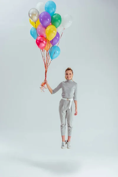 Красивая улыбающаяся женщина смотрит в камеру и прыгает в воздухе с пучком разноцветных воздушных шаров на сером фоне — стоковое фото