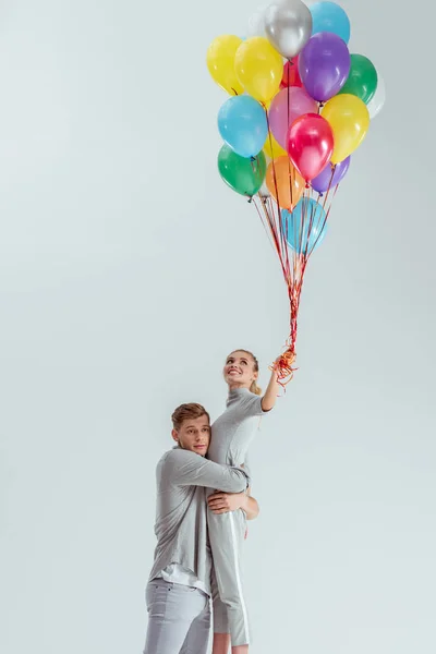 Hermosa pareja abrazándose mientras mujer celebración paquete de globos de colores aislados en gris - foto de stock