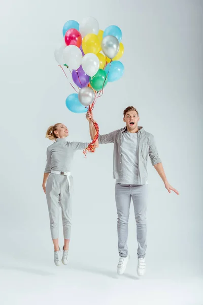 Взволнованная пара, прыгающая с пучком разноцветных воздушных шаров на сером фоне — стоковое фото