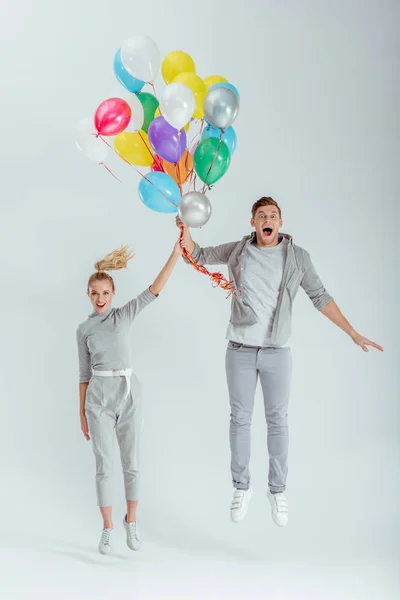 Aufgeregtes Paar blickt in die Kamera und springt mit einem Bündel bunter Luftballons auf grauem Hintergrund — Stockfoto