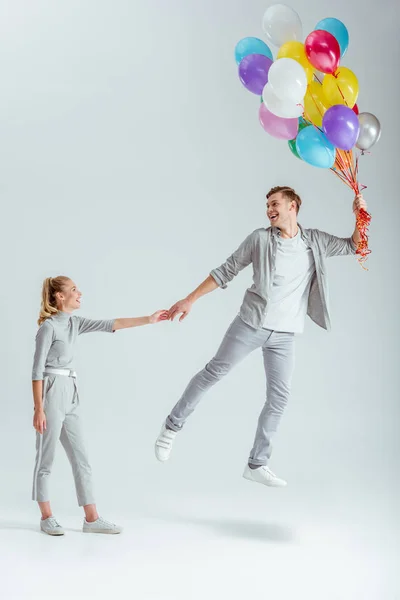Mujer en ropa gris cogida de la mano del hombre feliz saltando en el aire con un paquete de globos de colores sobre fondo gris - foto de stock