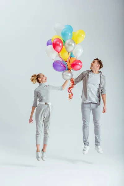 Aufgeregtes Paar in grauer Kleidung springt in der Luft mit einem Bündel bunter Luftballons auf grauem Hintergrund — Stockfoto