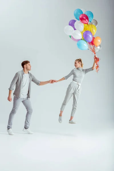 Hombre de la mano de la mujer saltando en el aire con un paquete de globos de colores sobre fondo gris - foto de stock